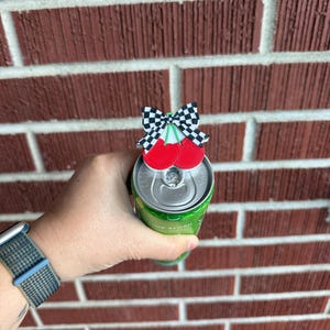 May include: A cherry-themed can topper with a black and white checkered bow and two red cherries. The topper is placed on top of a green can. The background is a red brick wall.