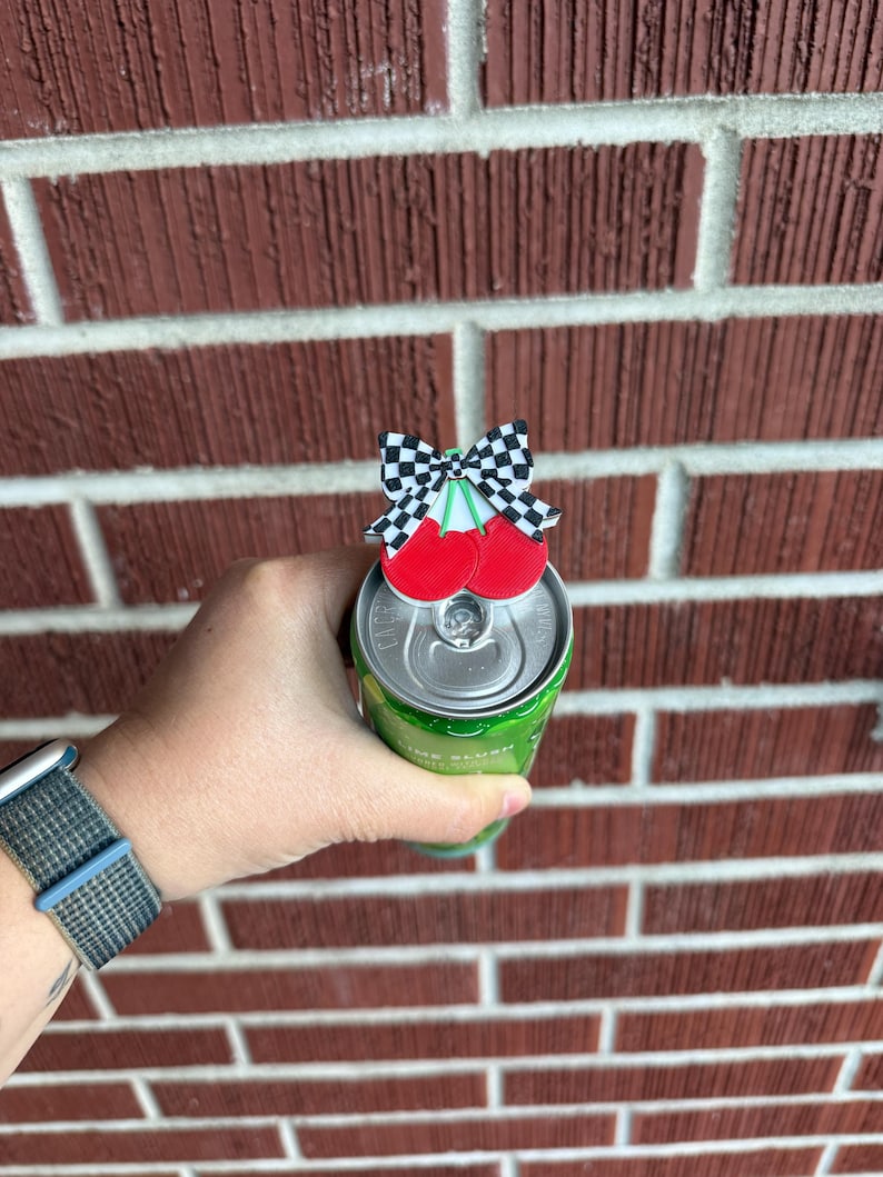 May include: A cherry-themed can topper with a black and white checkered bow and two red cherries. The topper is placed on top of a green can. The background is a red brick wall.