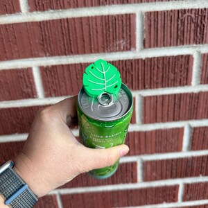 May include: A green leaf-shaped can topper is placed on top of a green and silver can. The can is labeled "Lime Slush". The can is held in a person's hand. The background is a red brick wall.