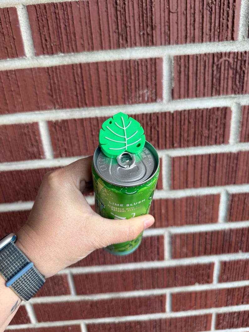 May include: A green leaf-shaped can topper is placed on top of a green and silver can. The can is labeled "Lime Slush". The can is held in a person's hand. The background is a red brick wall.