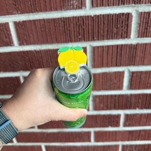 May include: A yellow lemon-shaped can topper with green leaves sits atop a green can. The can is held in a hand against a red brick wall. The can appears to contain a lime-flavored beverage.