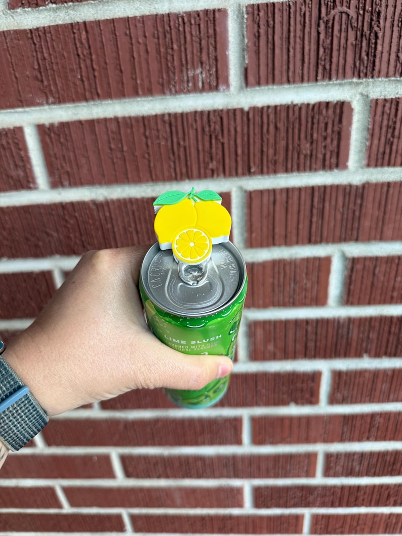 May include: A yellow lemon-shaped can topper with green leaves sits atop a green can. The can is held in a hand against a red brick wall. The can appears to contain a lime-flavored beverage.