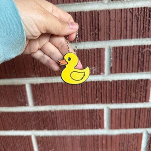 May include: A yellow duck keychain with a black outline and a small chain. The duck has an orange beak and pink accents. The keychain is held against a red brick wall.