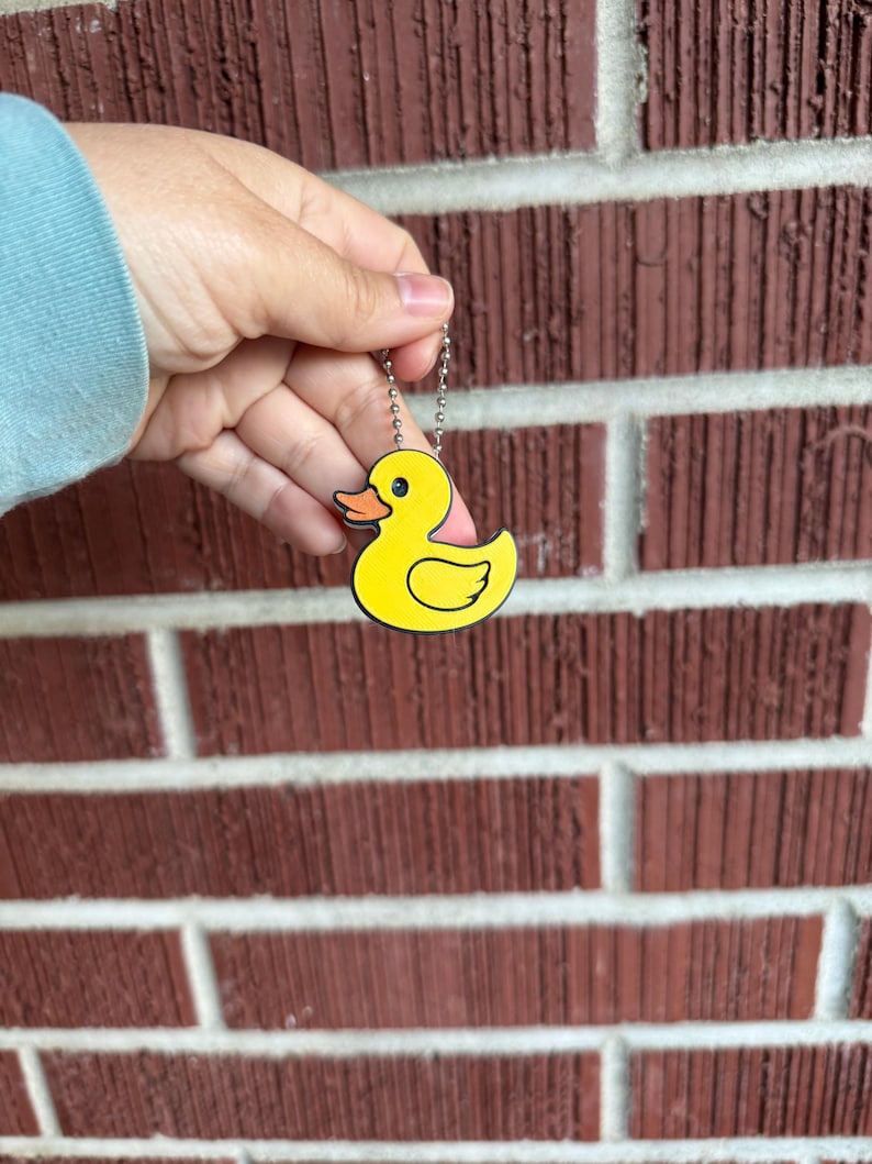 May include: A yellow duck keychain with a black outline and a small chain. The duck has an orange beak and pink accents. The keychain is held against a red brick wall.