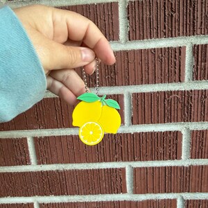 May include: A yellow lemon-shaped keychain with green leaves and a slice detail. The keychain is held against a red brick wall. The keychain is attached to a silver chain.