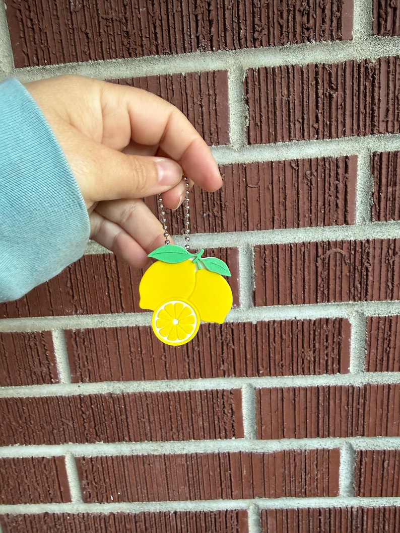 May include: A yellow lemon-shaped keychain with green leaves and a slice detail. The keychain is held against a red brick wall. The keychain is attached to a silver chain.