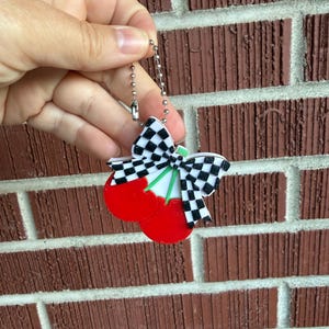 May include: A cherry-shaped keychain with a black and white checkered bow and green stems. The cherries are bright red. The keychain is held against a brick wall.