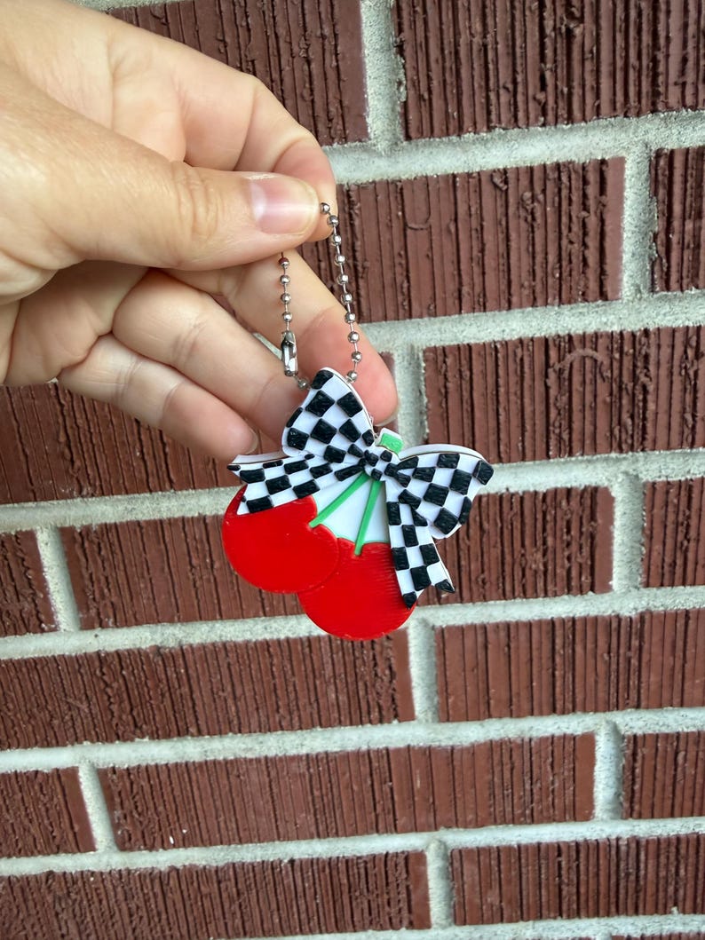 May include: A cherry-shaped keychain with a black and white checkered bow and green stems. The cherries are bright red. The keychain is held against a brick wall.