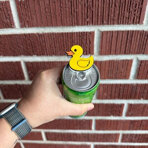 May include: A yellow duck-shaped can topper with an orange beak is placed on top of a green can. The can is held in a hand, with a brick wall in the background. The watch on the wrist has a blue and gray band.