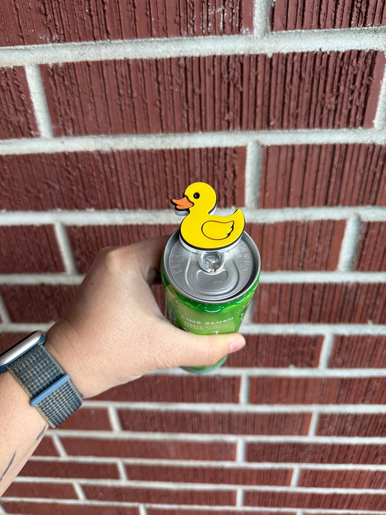 May include: A yellow duck-shaped can topper with an orange beak is placed on top of a green can. The can is held in a hand, with a brick wall in the background. The watch on the wrist has a blue and gray band.