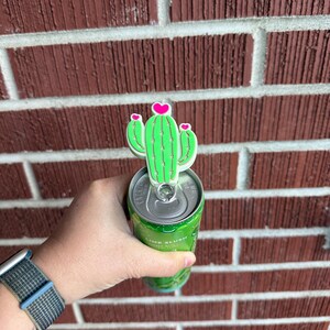 May include: A green cactus-shaped can topper with pink heart accents is placed on top of a green can. The can is held in a person's hand. The background is a red brick wall.