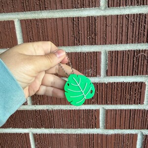 May include: A bright green leaf-shaped keychain with white vein details. The keychain is attached to a silver ball chain. The leaf has two small holes and is held against a brick wall background.