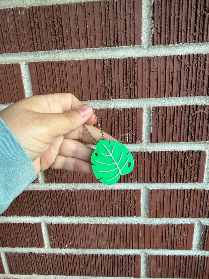 May include: A bright green leaf-shaped keychain with white vein details. The keychain is attached to a silver ball chain. The leaf has two small holes and is held against a brick wall background.