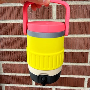 Puede incluir: Una jarra de agua multicolor con asa y tapa rosa, banda gris, sección media amarilla, base blanquecina y fondo negro con boquilla. La jarra se sostiene contra una pared de ladrillos rojos.