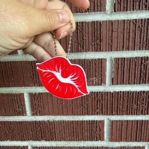 May include: A red and white lips-shaped keychain with a silver chain. The keychain is held in front of a brown brick wall.
