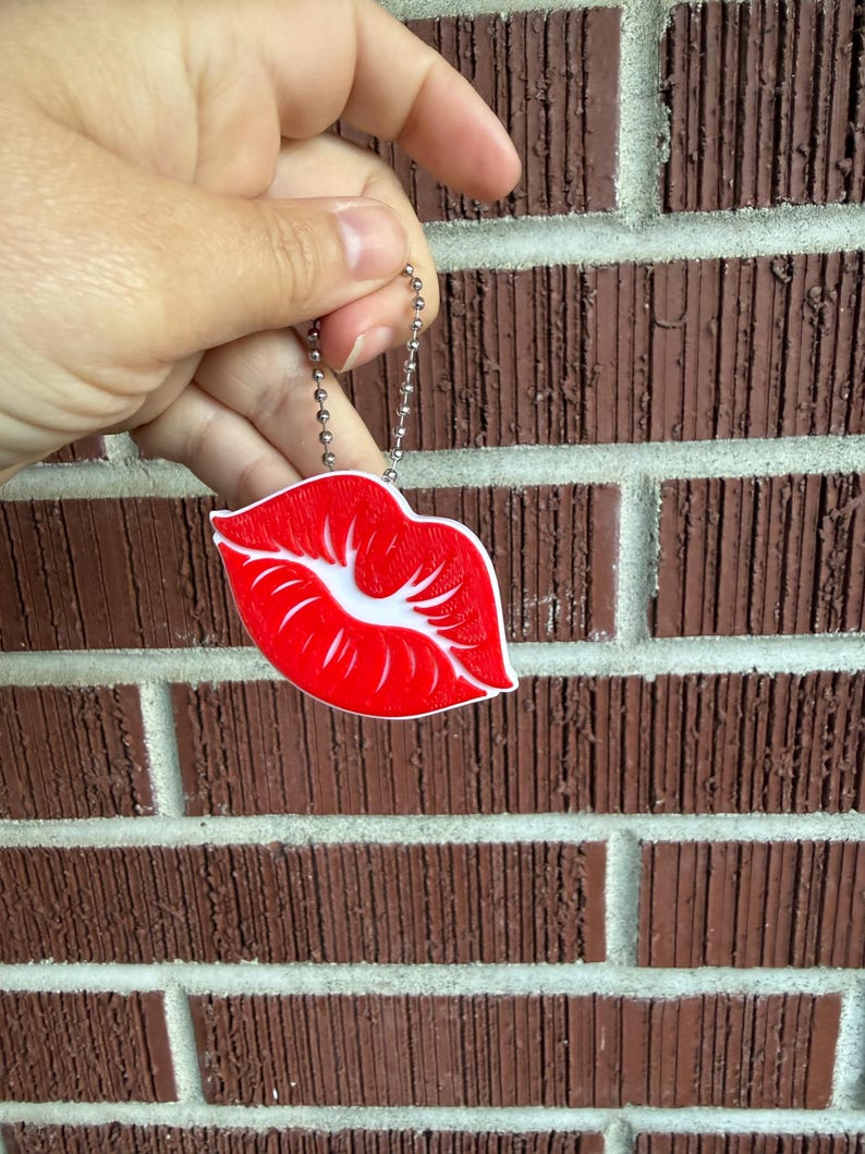 May include: A red and white lips-shaped keychain with a silver chain. The keychain is held in front of a brown brick wall.