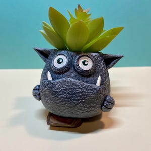 May include: A gray ceramic planter shaped like a monster with large eyes and white teeth. The planter has a green succulent plant growing out of the top.