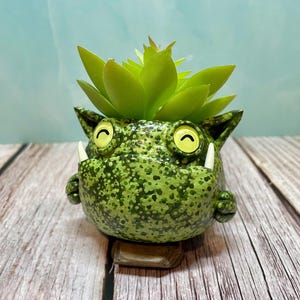 May include: A green ceramic planter shaped like a cartoon monster with a toothy grin and large eyes. The planter is decorated with a speckled green pattern and has a small succulent plant growing out of the top.