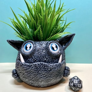 May include: A gray ceramic planter shaped like a monster with blue eyes and white teeth. The planter has a green plant growing out of the top. A black and white dice with the numbers 14, 13, 12, 11, 10, and 9 is sitting next to the planter.