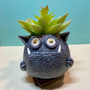 May include: A gray ceramic planter shaped like a monster with large eyes and fangs. The planter has a green succulent plant growing out of the top.