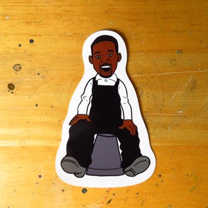 May include: A cartoon sticker of a Black man wearing overalls and a white shirt, sitting on a grey bucket. He is smiling and has his mouth open.
