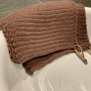Può includere: Un panno da bagno di maglia marrone con lacci in pelle. Il panno da bagno è piegato e posizionato in una ciotola di ceramica bianca.