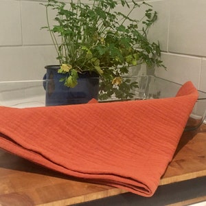 Può includere: Un asciugamano da cucina arancione piegato su un tagliere di legno accanto a una pirofila di vetro e un vaso di ceramica blu con una pianta verde.