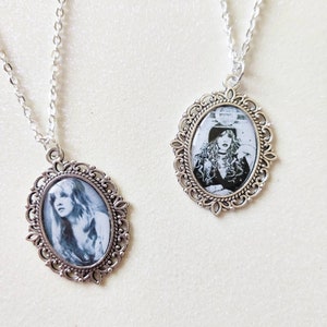 May include: Two silver-toned necklaces with oval pendants featuring black and white portraits. The pendants are framed with ornate, filigree-style borders. The chains are delicate and the background is a light, neutral color.