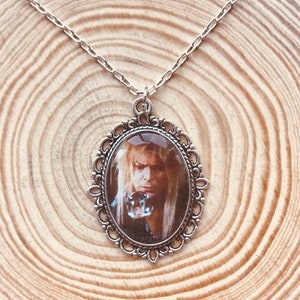 May include: Silver-toned necklace featuring an oval pendant with a portrait of a person with long blonde hair. The pendant is framed by an ornate, filigree-style border. The chain is delicate and the pendant is suspended from a small loop.