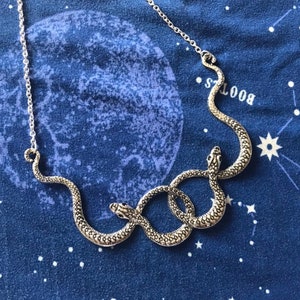 May include: Silver-toned snake necklace with three intertwined snakes. The necklace is on a dark blue fabric with a celestial pattern of stars and a large moon. The snakes have detailed scales.