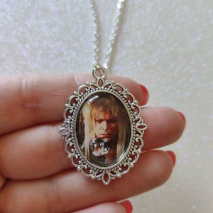 May include: Silver-toned pendant necklace featuring an oval-shaped pendant with a portrait of a person with long blonde hair. The pendant has a decorative filigree border and hangs from a delicate chain. The image is set against a light background.