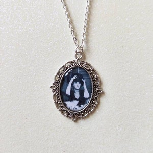 May include: Silver-toned necklace with an oval pendant featuring a black and white image of a person. The pendant is framed by an ornate, filigree-style border. The chain is delicate and hangs from the pendant.
