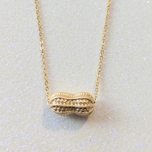 May include: A gold-coloured peanut pendant necklace. The peanut charm features textured surfaces and hangs from a delicate gold chain. The necklace is displayed against a sparkly, white background.
