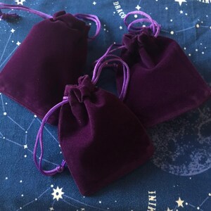 May include: Three deep purple velvet drawstring pouches with matching purple cords. The pouches are arranged on a dark blue fabric with a white celestial pattern. The pouches are closed, with the cords cinched at the top.