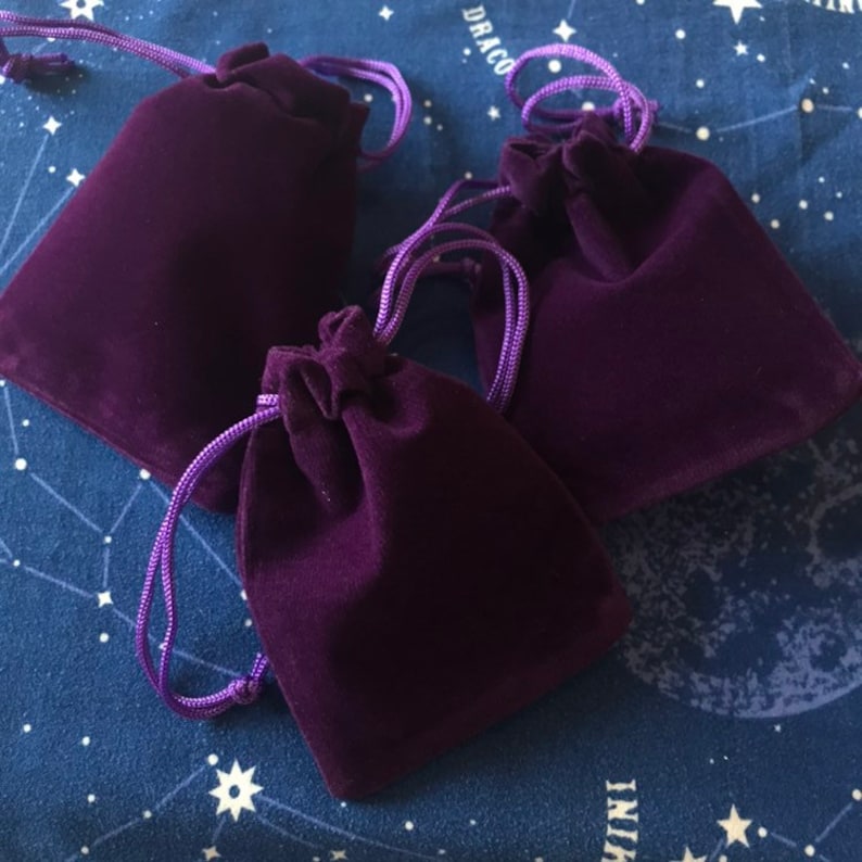 May include: Three deep purple velvet drawstring pouches with matching purple cords. The pouches are arranged on a dark blue fabric with a white celestial pattern. The pouches are closed, with the cords cinched at the top.