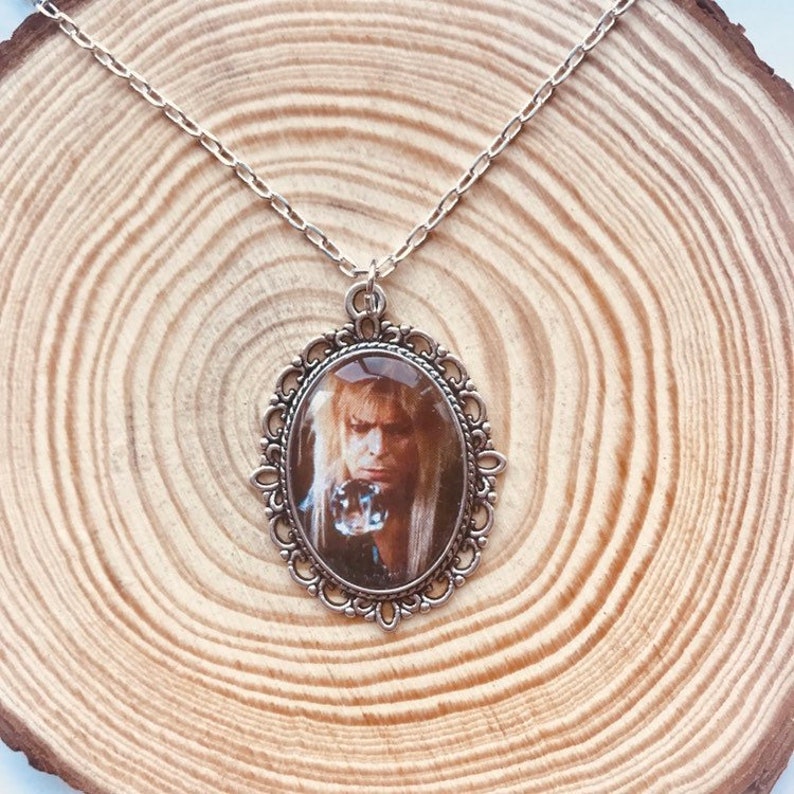 May include: Silver-toned necklace featuring an oval pendant with a portrait of a person with long blonde hair. The pendant is framed by an ornate, filigree-style border. The chain is delicate and the pendant is suspended from a small loop.