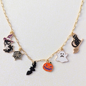 May include: Gold-coloured chain necklace with Halloween-themed charms. Charms include a black cat in a witch's hat, a spider web, a bat, a jack-o'-lantern, a sad ghost, and a black cat on a broom. The charms are black, orange, purple, and white.