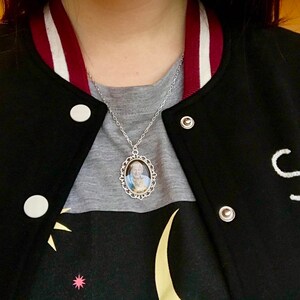May include: Silver-toned necklace with an oval pendant featuring a portrait. The pendant is framed with a decorative filigree design. The necklace is worn over a gray shirt and a black varsity jacket with white lettering.