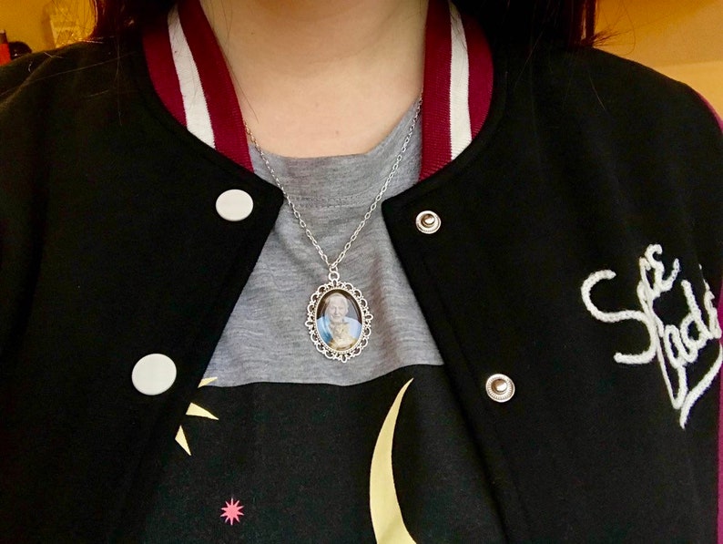 May include: Silver-toned necklace with an oval pendant featuring a portrait. The pendant is framed with a decorative filigree design. The necklace is worn over a gray shirt and a black varsity jacket with white lettering.