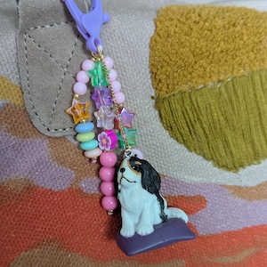 May include: A decorative keychain with a small dog figurine in black and white, resting on a purple cushion. The keychain has a purple clasp, star-shaped beads, and pink and pastel beads, creating a fun accessory.