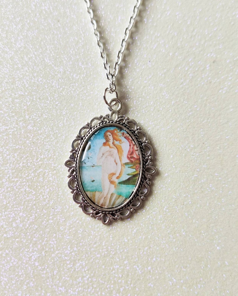 A silver oval jewellery charm, with a photo of Botticelli's Birth of Venus painting. Attached to a silver chain, against a creamy, sparkly background.