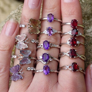 May include: A hand wearing multiple silver rings with various gemstones. The rings feature butterfly designs, oval-shaped amethyst stones, and round red garnet stones.