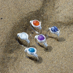 Könnte beinhalten: Sechs drahtgewickelte Ringe mit verschiedenen farbigen Edelsteinen. Die Ringe liegen auf einer sandigen Oberfläche.