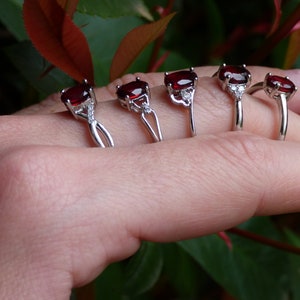 May include: Five silver rings with red gemstones and small clear stones. The rings are arranged in a row on a hand, with the largest ring on the pinky finger and the smallest ring on the thumb.