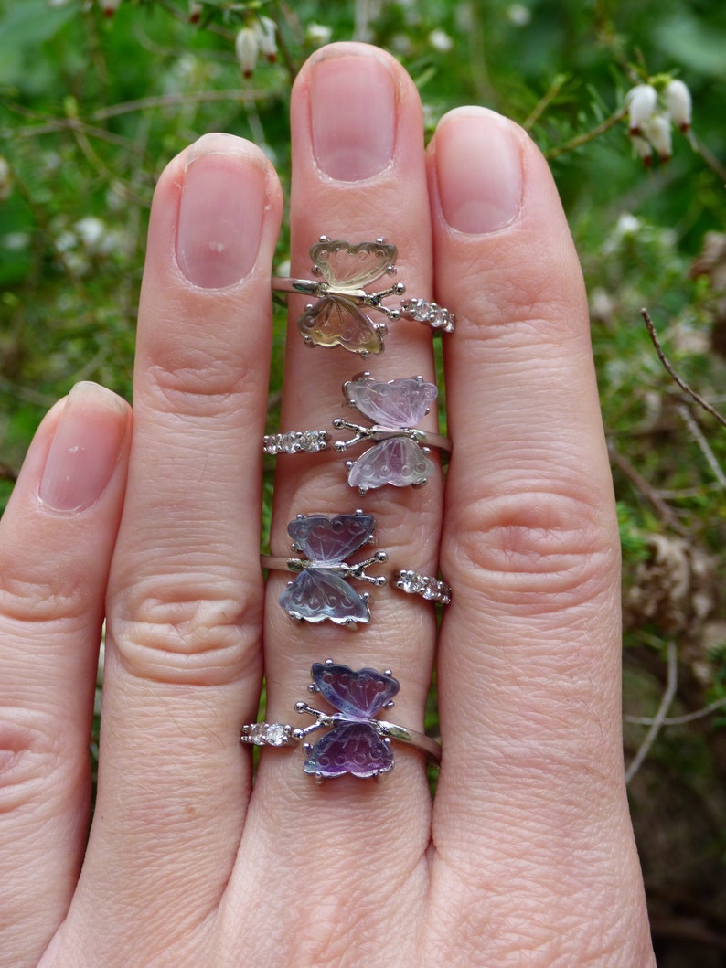 May include: Five silver rings with butterfly-shaped gemstones. The butterflies are iridescent and have different colors, including purple, pink, and green.