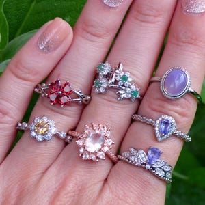 Könnte beinhalten: Eine Hand trägt sechs Silberringe mit verschiedenen Edelsteinen. Die Ringe haben verschiedene Designs, darunter florale, geometrische und birnenförmige Steine. Die Steine sind eine Mischung aus Rosa, Lila, Grün und Rot.