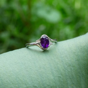 May include: A silver ring with a purple oval-shaped gemstone and two smaller clear gemstones. The ring is on a green leaf.