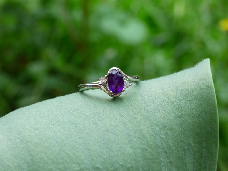 May include: A silver ring with a purple oval-shaped gemstone and two smaller clear gemstones. The ring is on a green leaf.