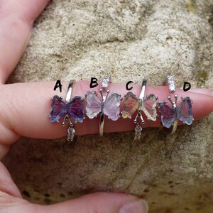 May include: Four silver butterfly rings with different colored butterfly wings. The rings are labeled A, B, C, and D. The butterfly wings are purple, pink, yellow, and blue.