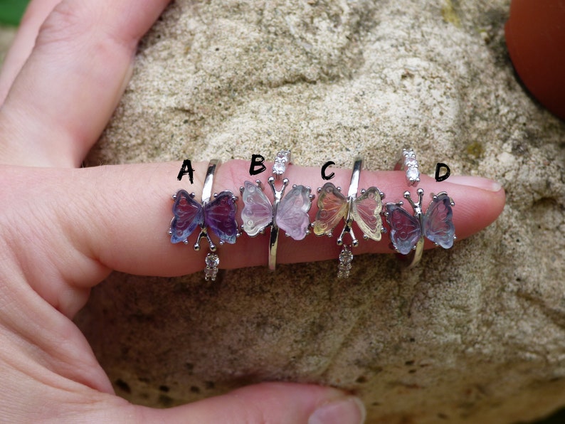 May include: Four silver butterfly rings with different colored butterfly wings. The rings are labeled A, B, C, and D. The butterfly wings are purple, pink, yellow, and blue.